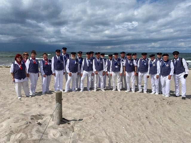 Rund 25 Sängerinnen und Sänger in blau-weißer Chorkleidung mit dunklen Schirmmützen stehen in einer langen Reihe am Ostseestrand, dahinter Meer und Wolken.