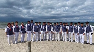 Rund 25 Sängerinnen und Sänger in blau-weißer Chorkleidung mit dunklen Schirmmützen stehen in einer langen Reihe am Ostseestrand, dahinter Meer und Wolken.