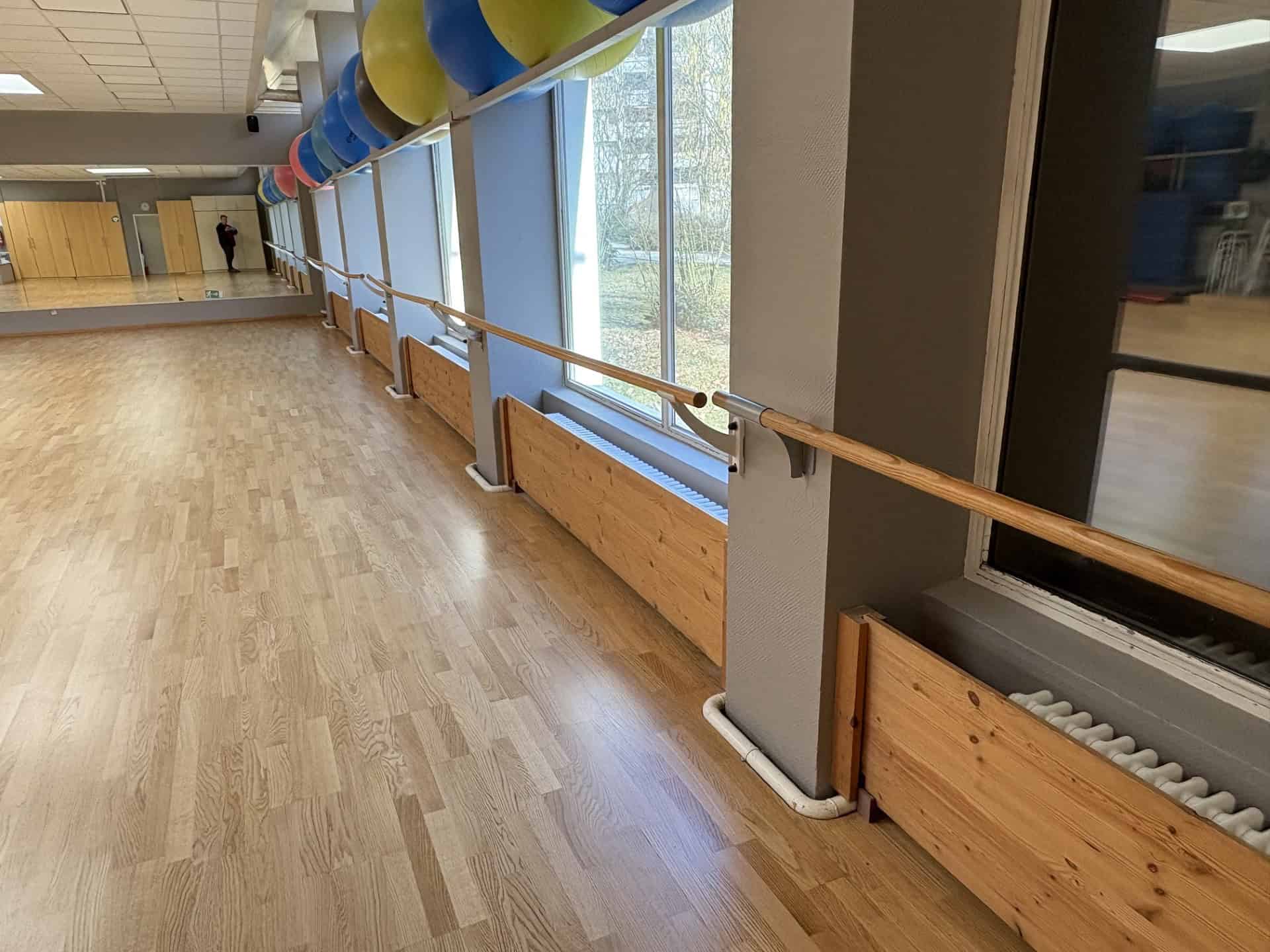 Heller Hallenraum mit Holzboden, Spiegelwand und Ballettstange; unter den Fensterfronten stehen Heizkörper in Holzverkleidungen, an der Decke hängen blaue und gelbe Luftballons.