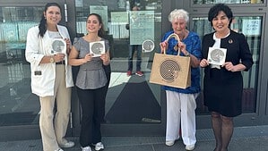 Vier Frauen stehen vor der Glastür eines Eingangs und halten Karten mit einem schwarz-weißen Spiralsymbol hoch; eine von ihnen trägt eine Jutetasche mit demselben Motiv.