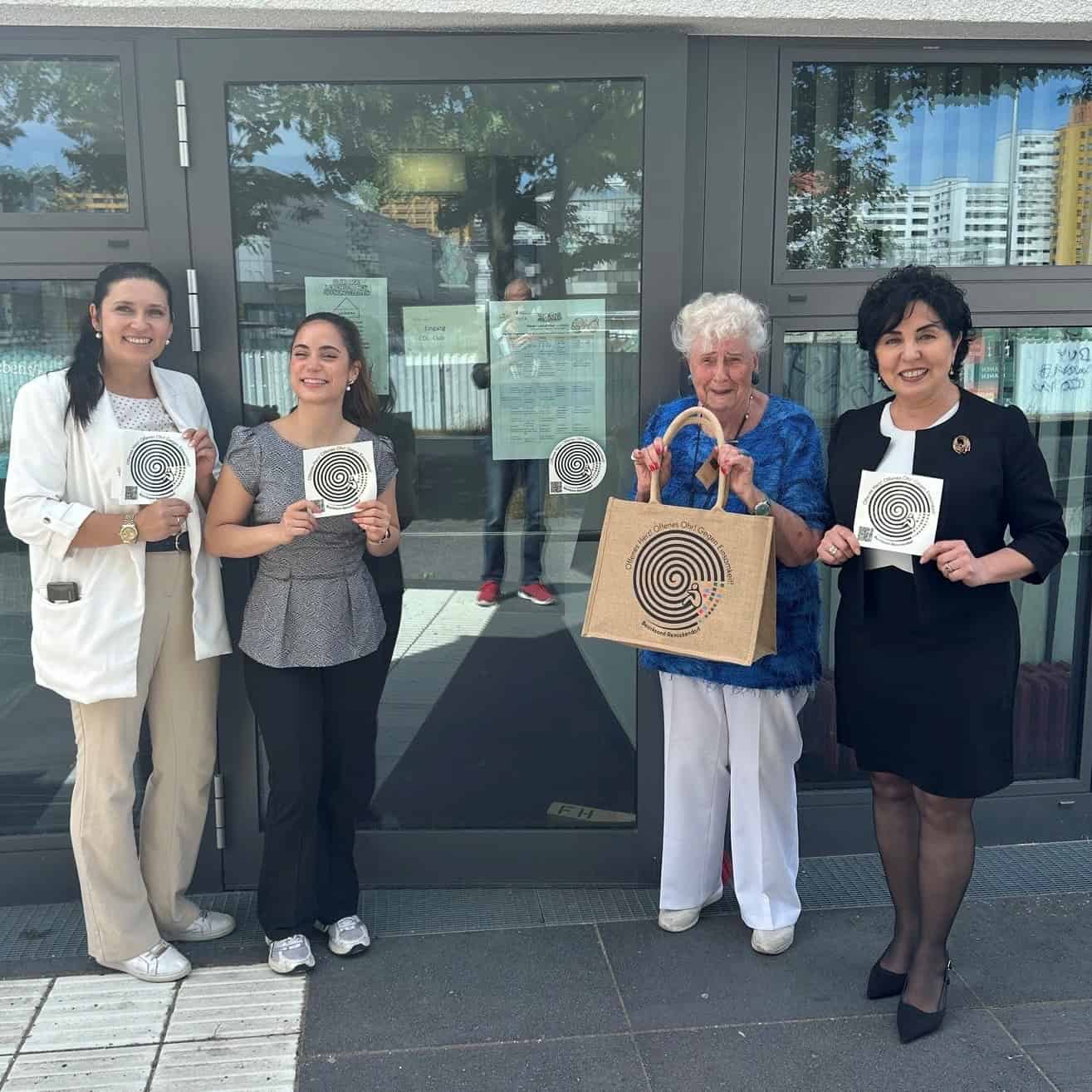Vier Frauen stehen vor der Glastür eines Eingangs und halten Karten mit einem schwarz-weißen Spiralsymbol hoch; eine von ihnen trägt eine Jutetasche mit demselben Motiv.