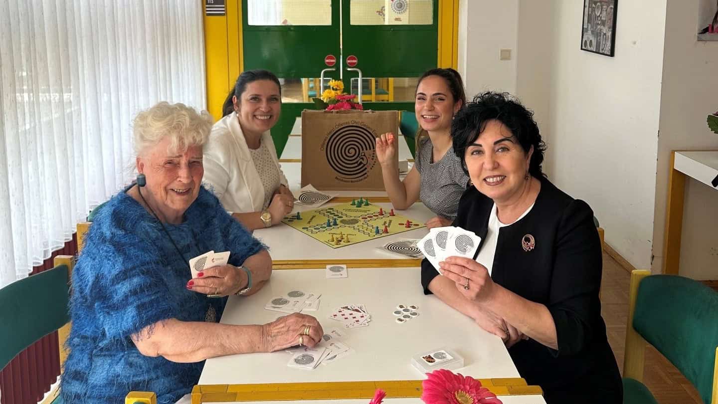Vier Frauen sitzen an einem langen weißen Tisch in einem Clubraum, halten Spielkarten in den Händen und lachen in die Kamera; auf dem Tisch stehen ein Brettspiel, Blumen und eine Jutetasche.
