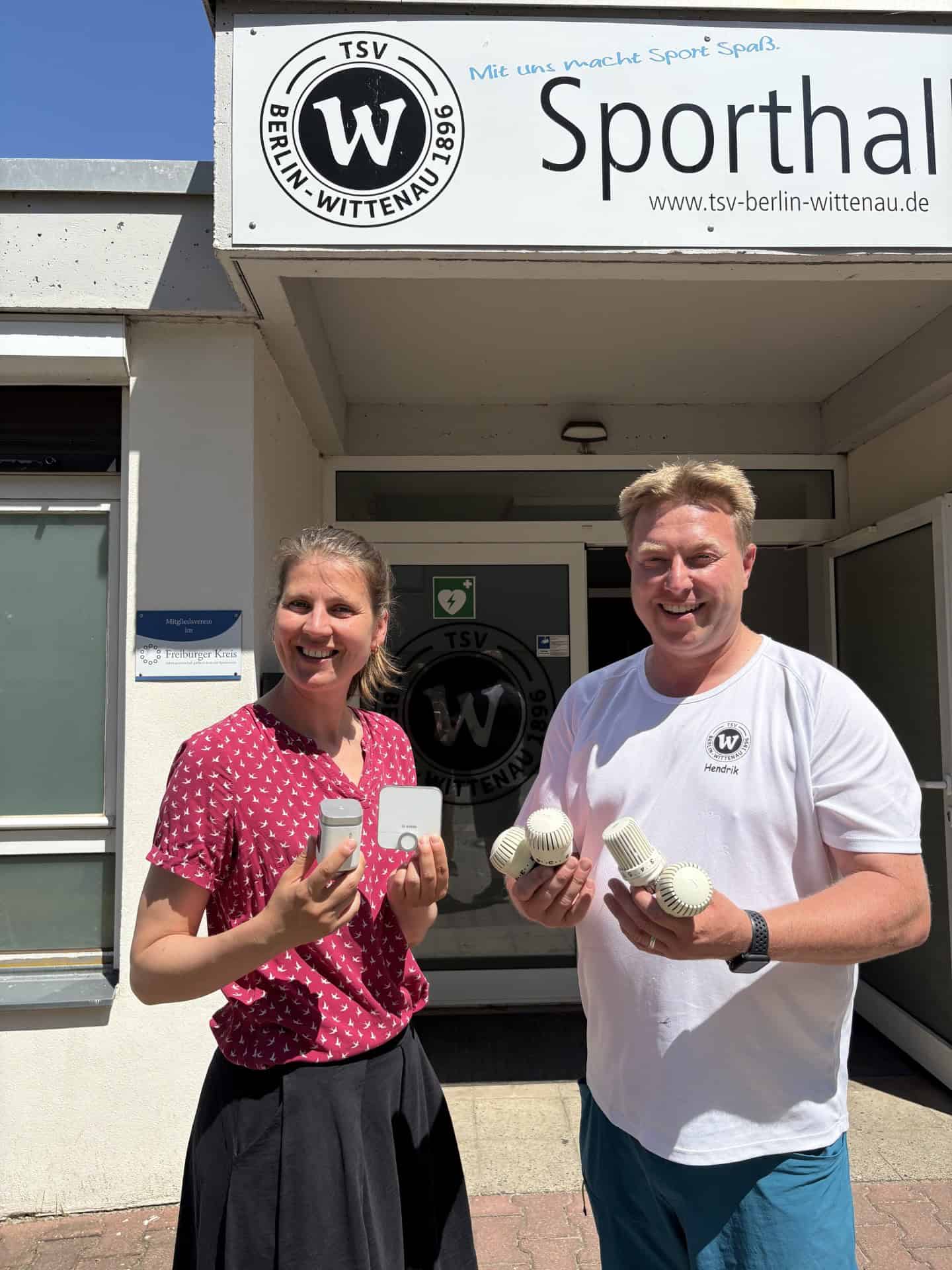 Eine Frau in rot gemusterter Bluse und ein Mann im weißen TSV-Shirt stehen vor dem Eingang der Vereinssporthalle und halten weiße digitale Thermostate sowie mehrere alte, geriffelte Thermostatköpfe in den Händen.
