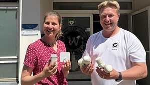Eine Frau in rot gemusterter Bluse und ein Mann im weißen TSV-Shirt stehen vor dem Eingang der Vereinssporthalle und halten weiße digitale Thermostate sowie mehrere alte, geriffelte Thermostatköpfe in den Händen.