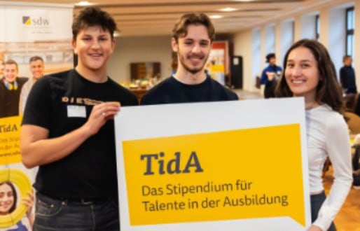 Drei junge Menschen halten ein Schild mit der Aufschrift TidA, das Stipendium für Talente in der Ausbildung