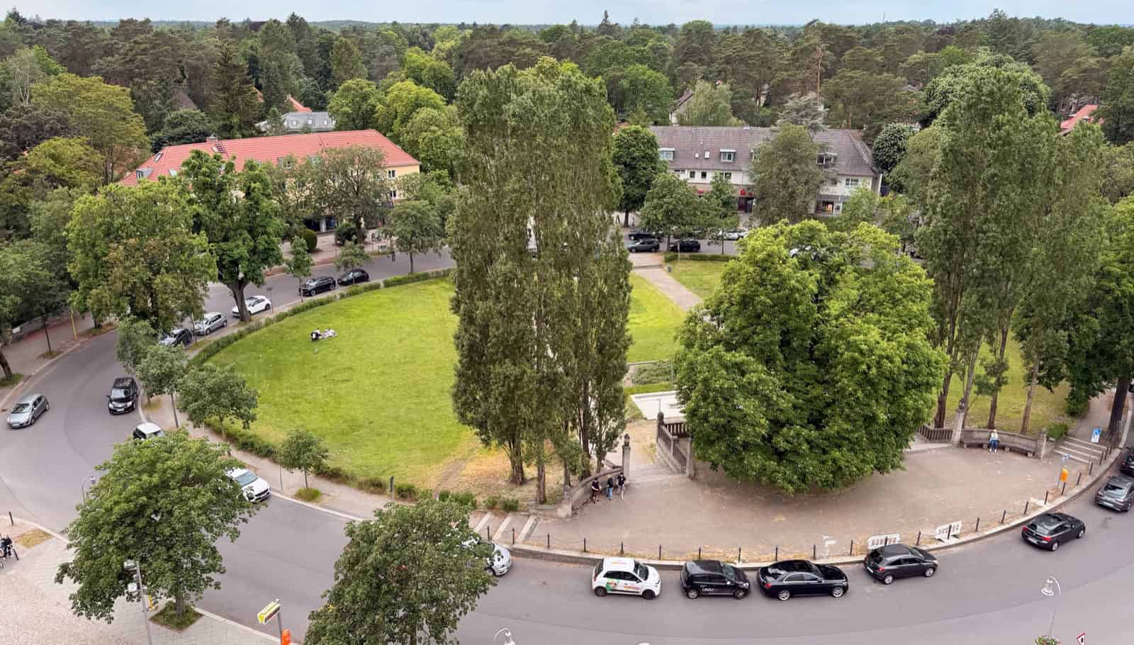 Luftaufnahme des Ludolfingerplatzes in Frohnau: eine ovale Rasenfläche mit hohen Pappeln in der Mitte, ringsum belaubte Straßenbäume, die Ringstraße mit parkenden Autos und dahinter Wohnhäuser am Waldrand.