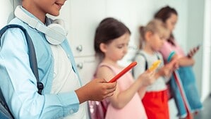 Vier Kinder mit Schulranzen stehen nebeneinander an einer hellen Garderobe und blicken auf ihre Smartphones.