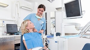 Eine Patientin sitzt im Behandlungsstuhl einer Zahnarztpraxis und blickt zu einer zahnmedizinischen Fachangestellten in blauer Berufskleidung auf.