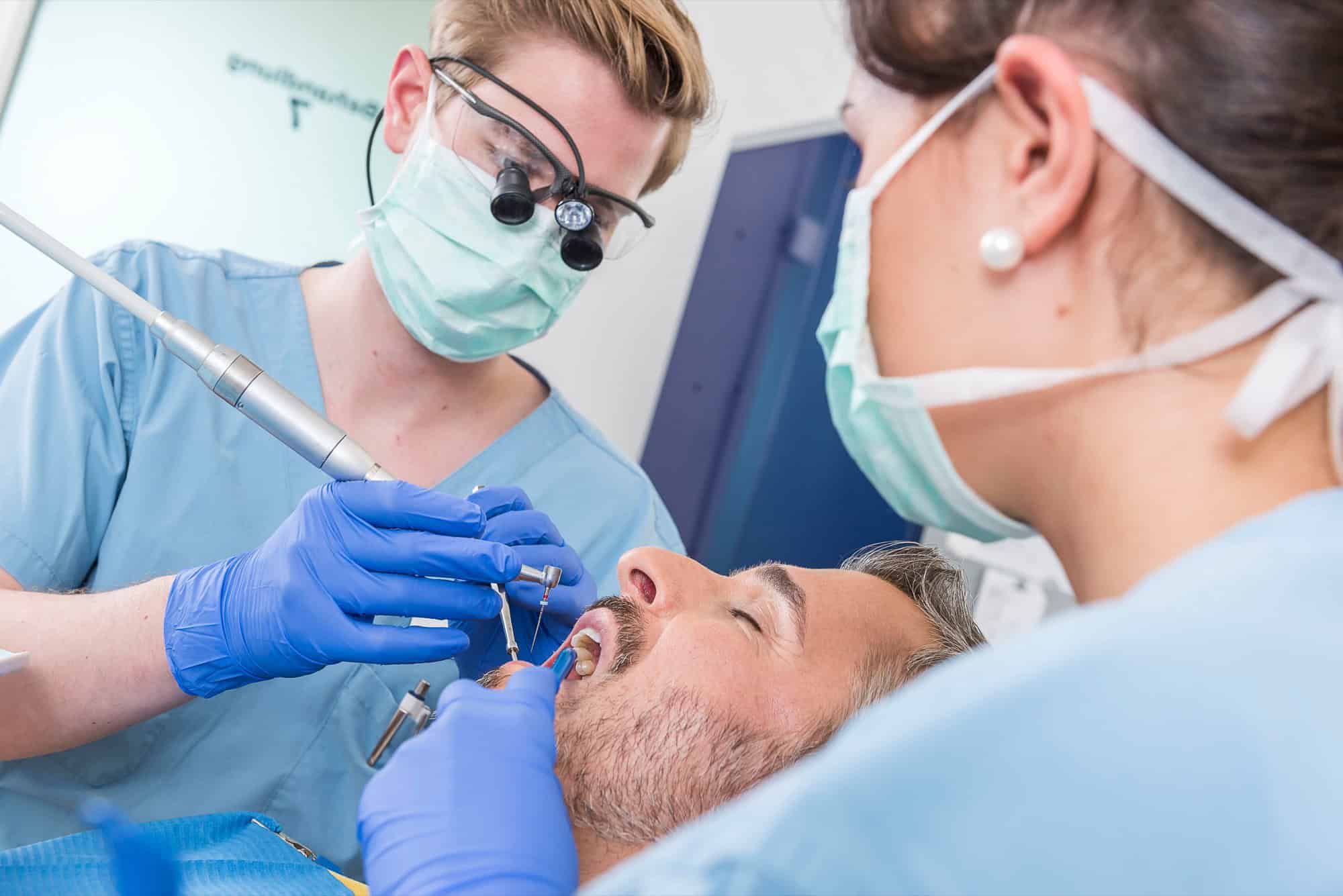 Ein Zahnarzt mit Lupenbrille und eine zahnmedizinische Fachangestellte behandeln einen Patienten, der im Behandlungsstuhl liegt.