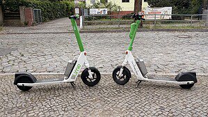 Zwei E-Roller stehen am Straßenrand. Im Hintergrund eine Kopfsteinpflasterstraße und ein Wohnhaus mit Plakaten am Zaun.