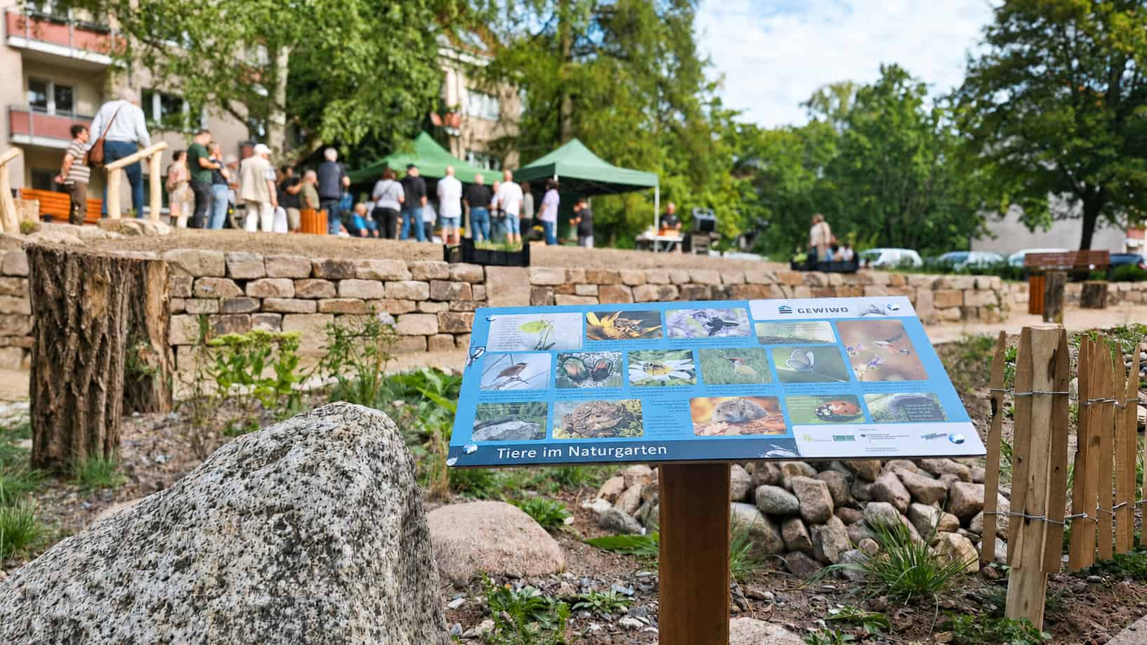 Informationsschild „Tiere im Naturgarten“ vor der neuen Grünfläche, dahinter Besucherinnen und Besucher der Eröffnung
