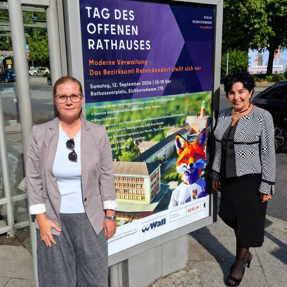 Zwei Frauen stehen links und rechts neben einer Plakatvitrine mit dem violetten Plakat zum Tag des offenen Rathauses, auf dem ein Fuchs im Anzug vor dem Rathaus abgebildet ist.