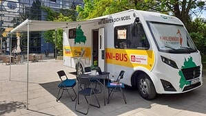 Weißer Beratungsbus mit gelber Beschriftung Boni-Bus und dem Schriftzug Familienbüro Reinickendorf steht mit ausgefahrener Markise, Tisch und Klappstühlen auf einem gepflasterten Vorplatz.