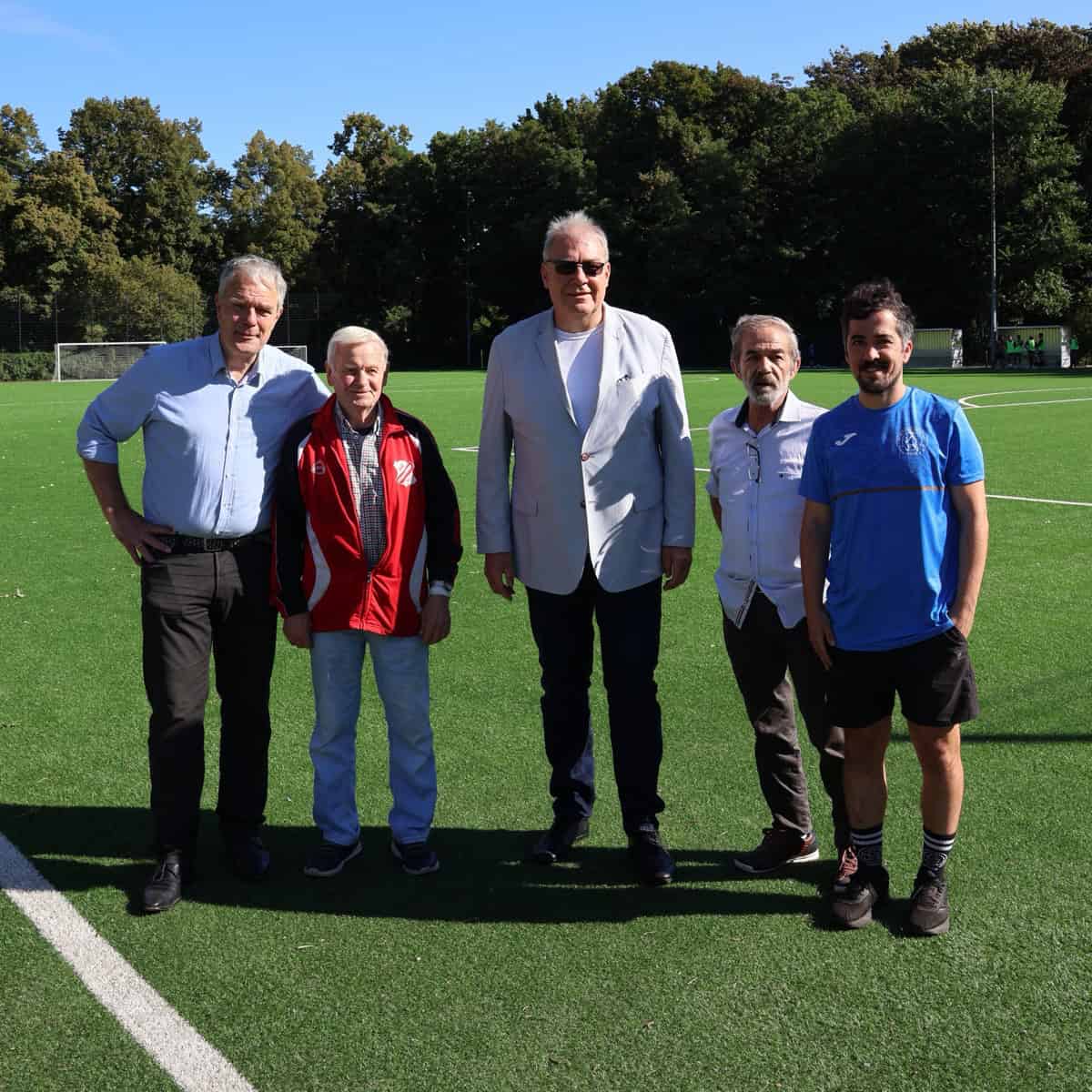 Fünf Männer stehen nebeneinander auf einem grünen Kunstrasenplatz, zwei von ihnen in Sportkleidung, dahinter Bäume und blauer Himmel.