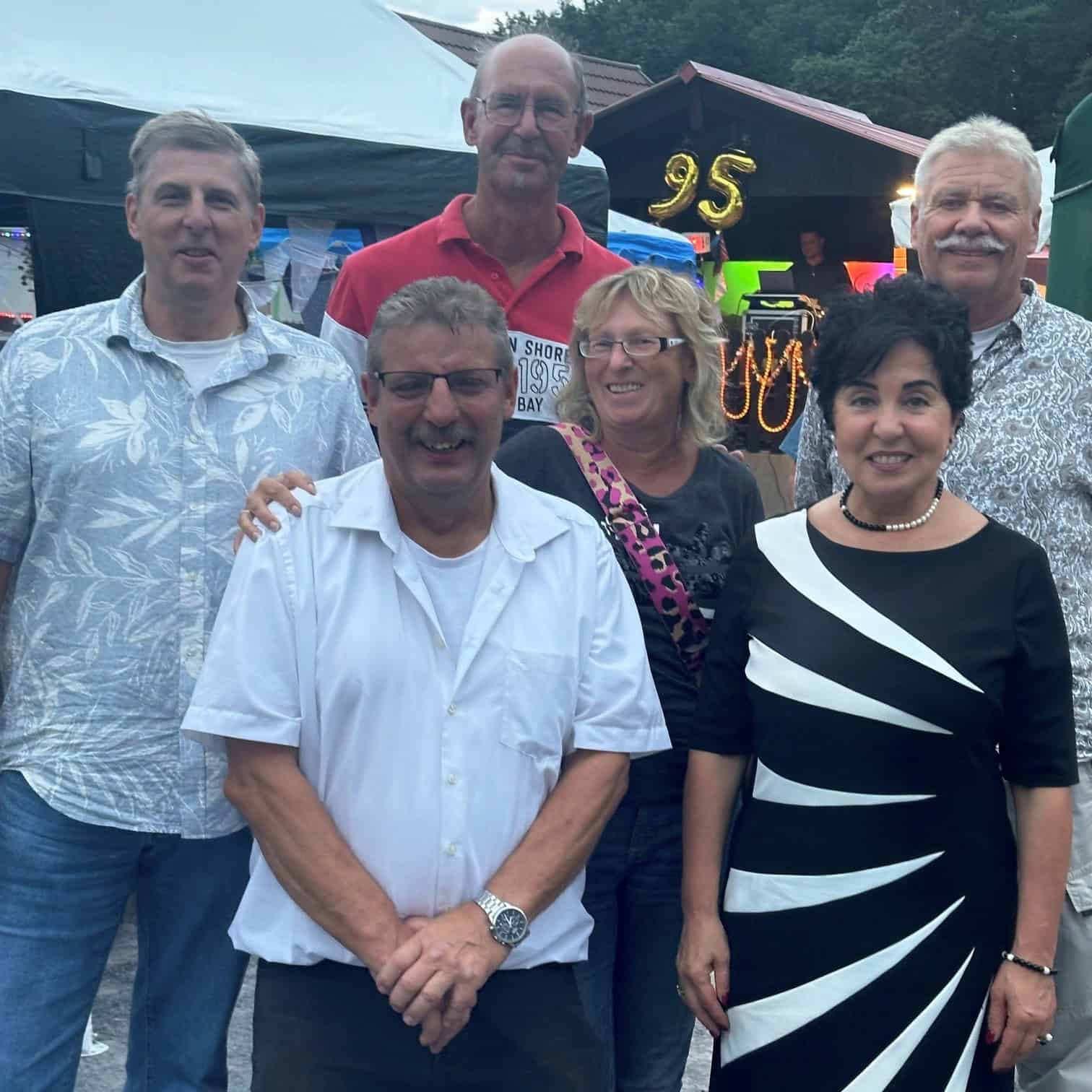 Sechs Personen stehen abends beim Jubiläumsfest dicht beieinander, im Hintergrund leuchtet eine Ballonzahl 95.