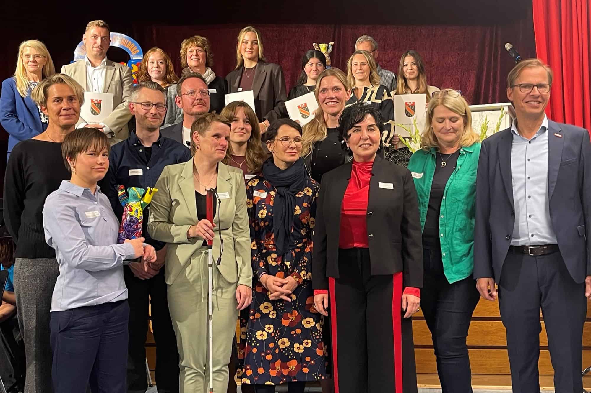Eine große Gruppe von Frauen und Männern steht mit Urkunden nebeneinander für ein Gruppenbild vor einem roten Vorhang.