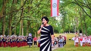 Bezirksbürgermeisterin Emine Demirbüken-Wegner spricht mit Mikrofon vor Bäumen, dahinter uniformierte Spielleute und Gardegruppen.