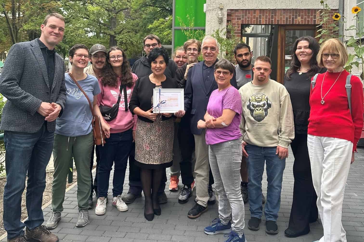 Bezirksbürgermeisterin Emine Demirbüken-Wegner hält vor dem Gebäude der Mosaik-Berlin GmbH einen gerahmten Spendenscheck, umringt von Beschäftigten und Gästen.