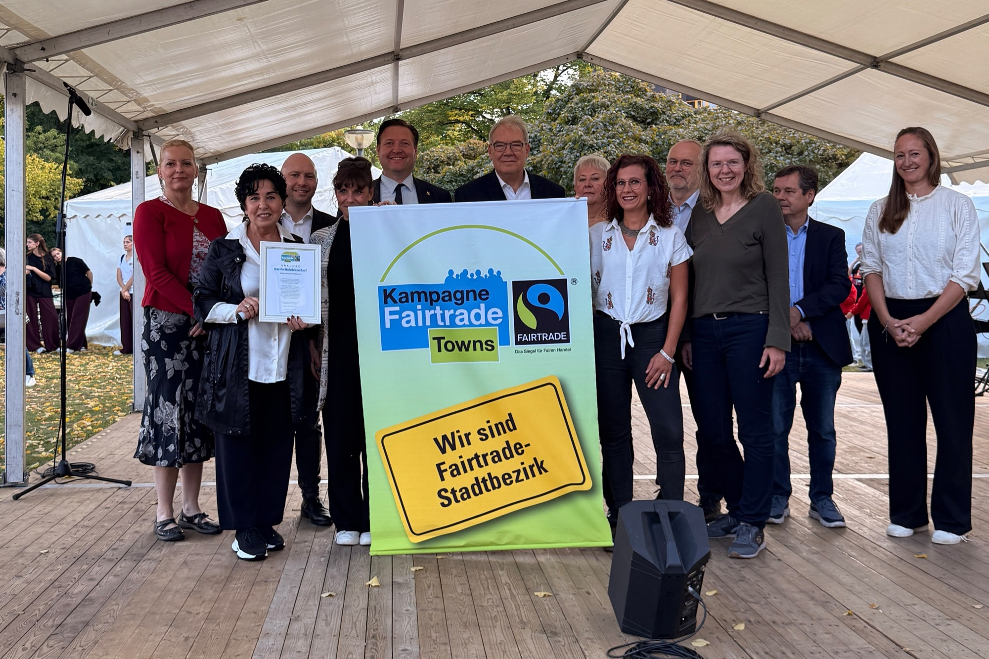 Zwölf Personen stehen auf einer Bühne hinter einem Plakat der Kampagne Fairtrade Towns mit der Aufschrift „Wir sind Fairtrade-Stadtbezirk“, eine Frau hält eine Urkunde