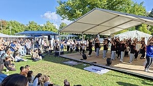 Kinder in schwarzen T-Shirts tanzen auf einer überdachten Bühne, davor sitzt und steht das Publikum auf dem Rasen