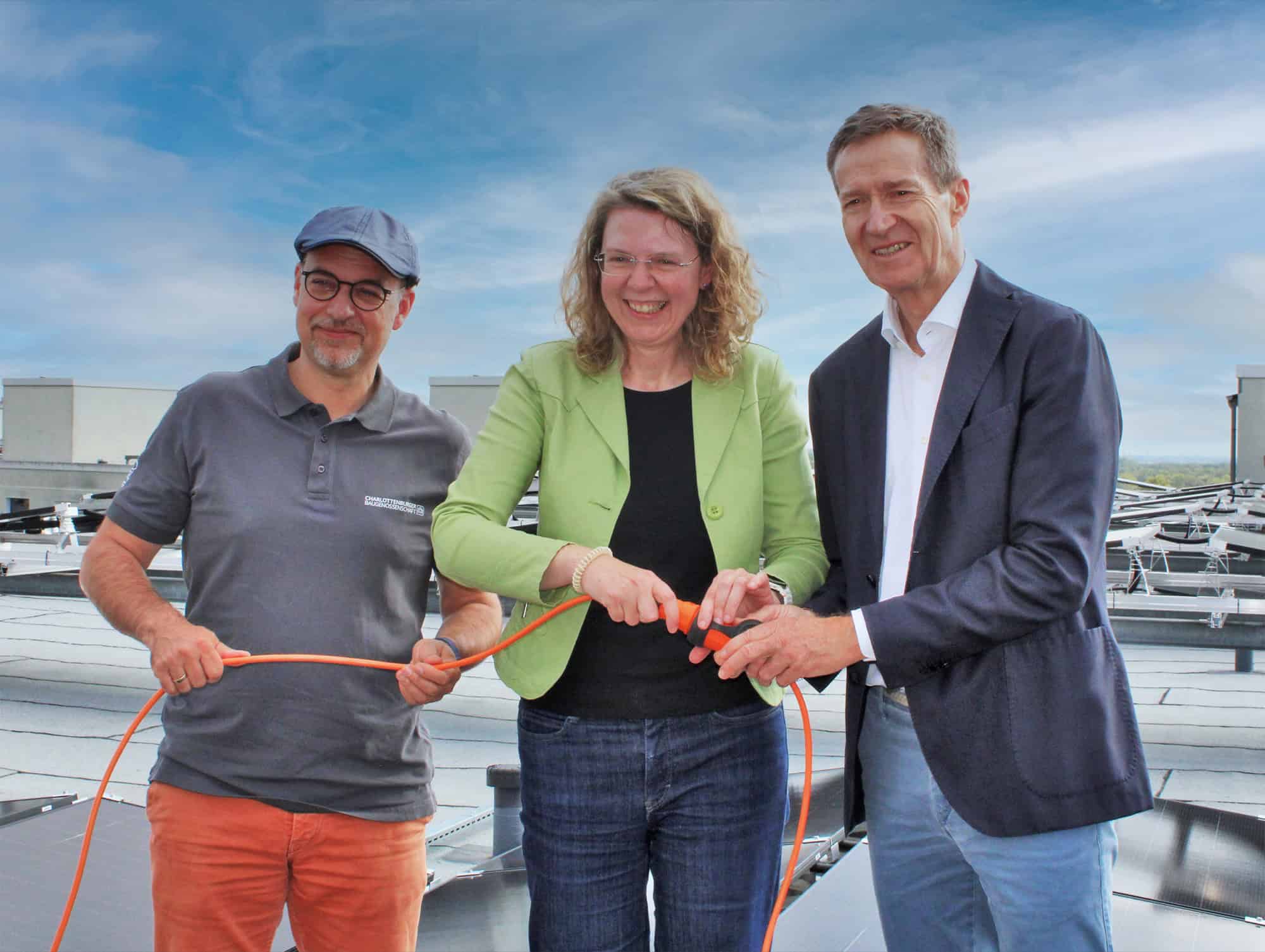 Drei Personen stehen auf einem Flachdach zwischen Solarmodulen und halten ein orangefarbenes Kabel.