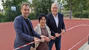 Drei Personen stehen auf einem roten Sportbelag und halten ein rot-weißes Absperrband, das durchtrennt wird.