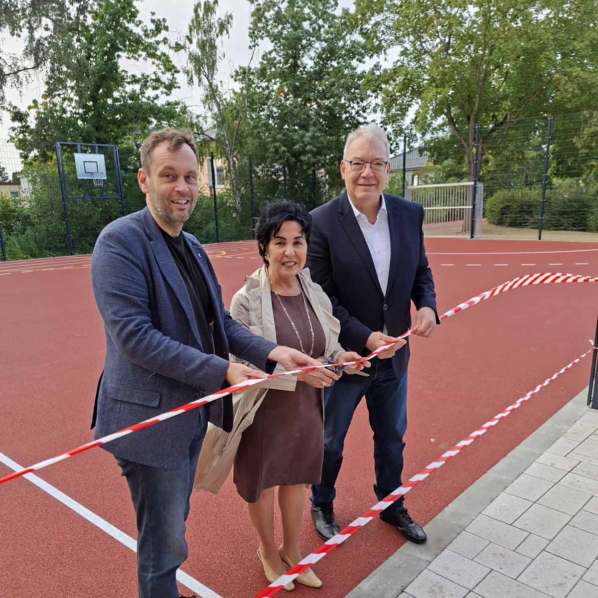 Drei Personen stehen auf einem roten Sportbelag und halten ein rot-weißes Absperrband, das durchtrennt wird.