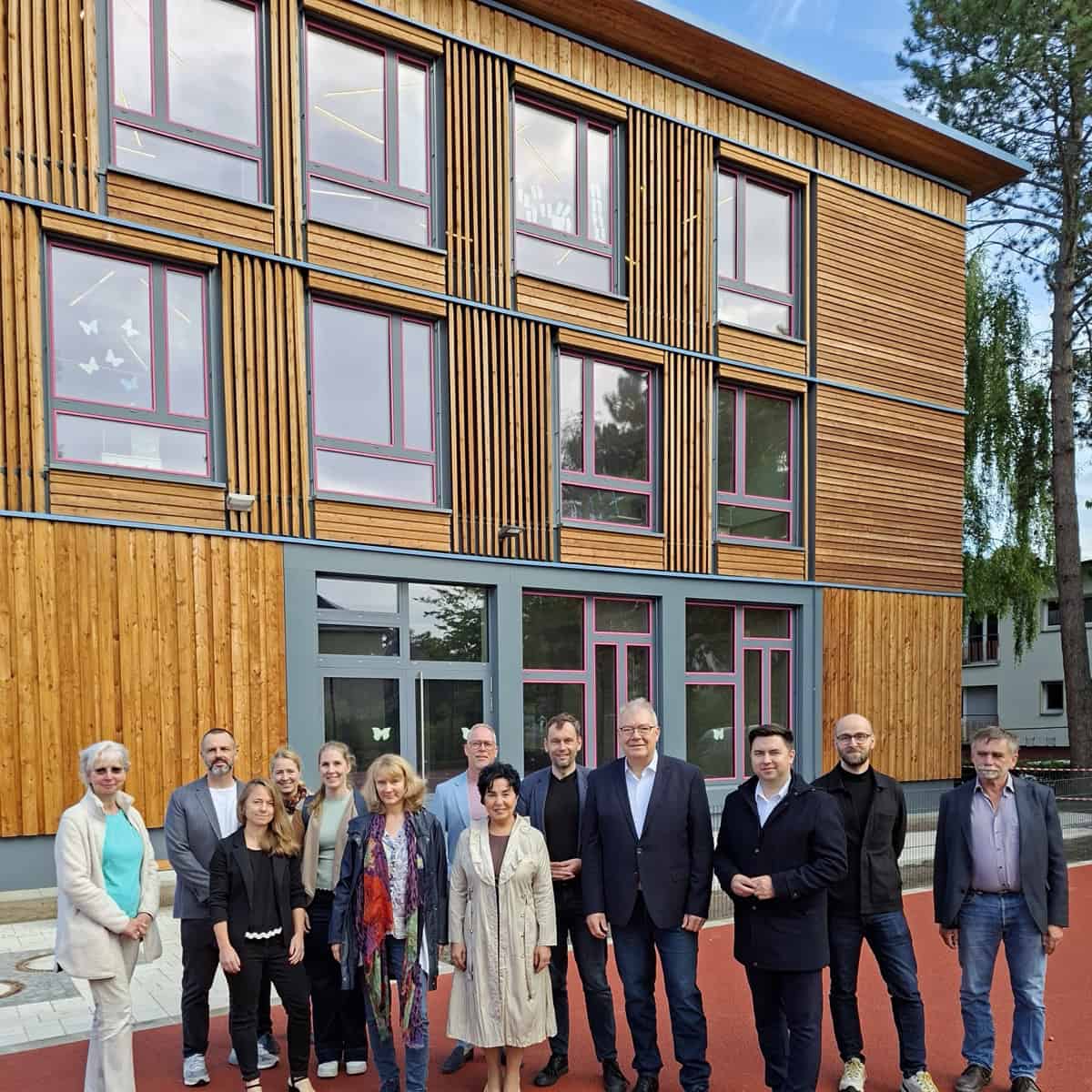 Eine Gruppe von rund vierzehn Erwachsenen steht auf einer roten Spielfläche vor einem Schulgebäude mit Holzfassade.