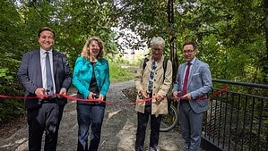 Vier Personen durchschneiden auf einem frisch angelegten Weg im Grünen gemeinsam ein rotes Band, rechts ist das Geländer einer Brücke zu sehen.