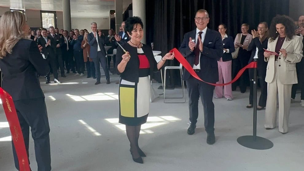 Eine Frau durchschneidet in einer hellen Halle mit Betondecke ein rotes Band, um sie herum applaudieren zahlreiche Gäste.