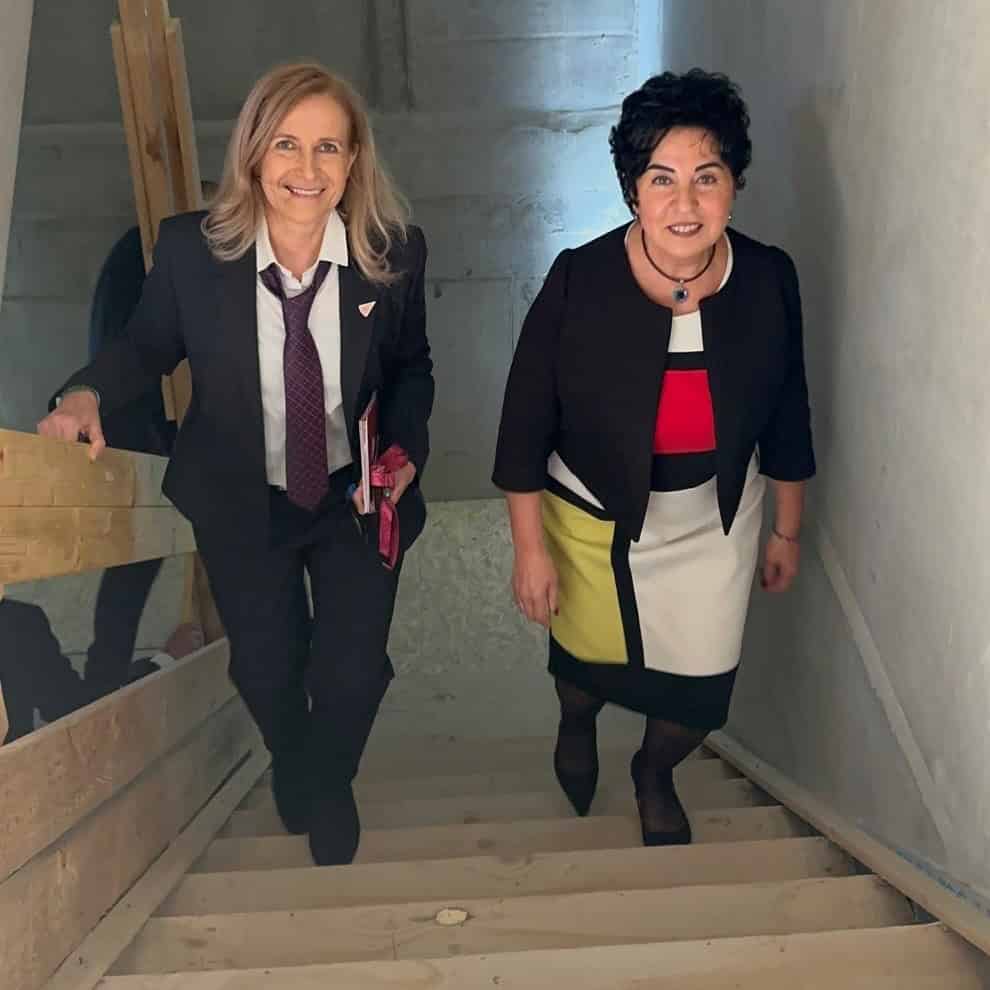 Zwei Frauen steigen lächelnd eine Rohbautreppe mit Holzstufen empor.