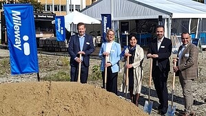 Fünf Personen in Geschäftskleidung stehen mit Spaten hinter einem Sandhaufen, links und rechts blaue Mileway-Fahnen, im Hintergrund ein Festzelt und ein Bürogebäude.