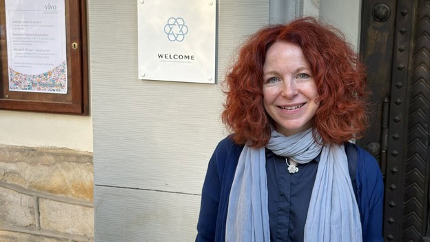Eine Frau steht lächelnd neben einem Schild mit der Aufschrift Welcome am Portal der Dorfkirche Alt-Tegel