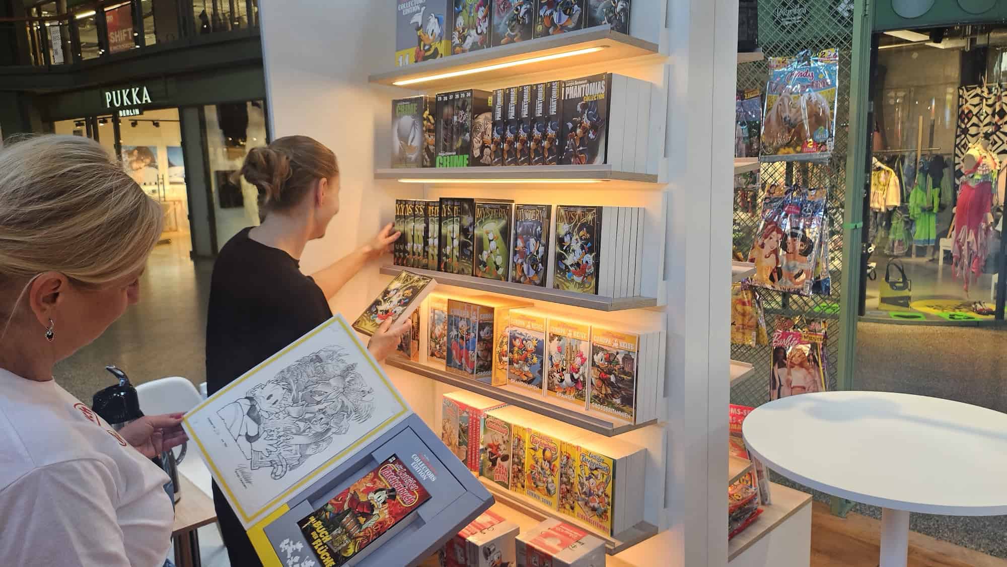 Zwei Frauen stehen vor beleuchteten Regalen mit Disney-Comicbänden im COMIC POP UP-Store