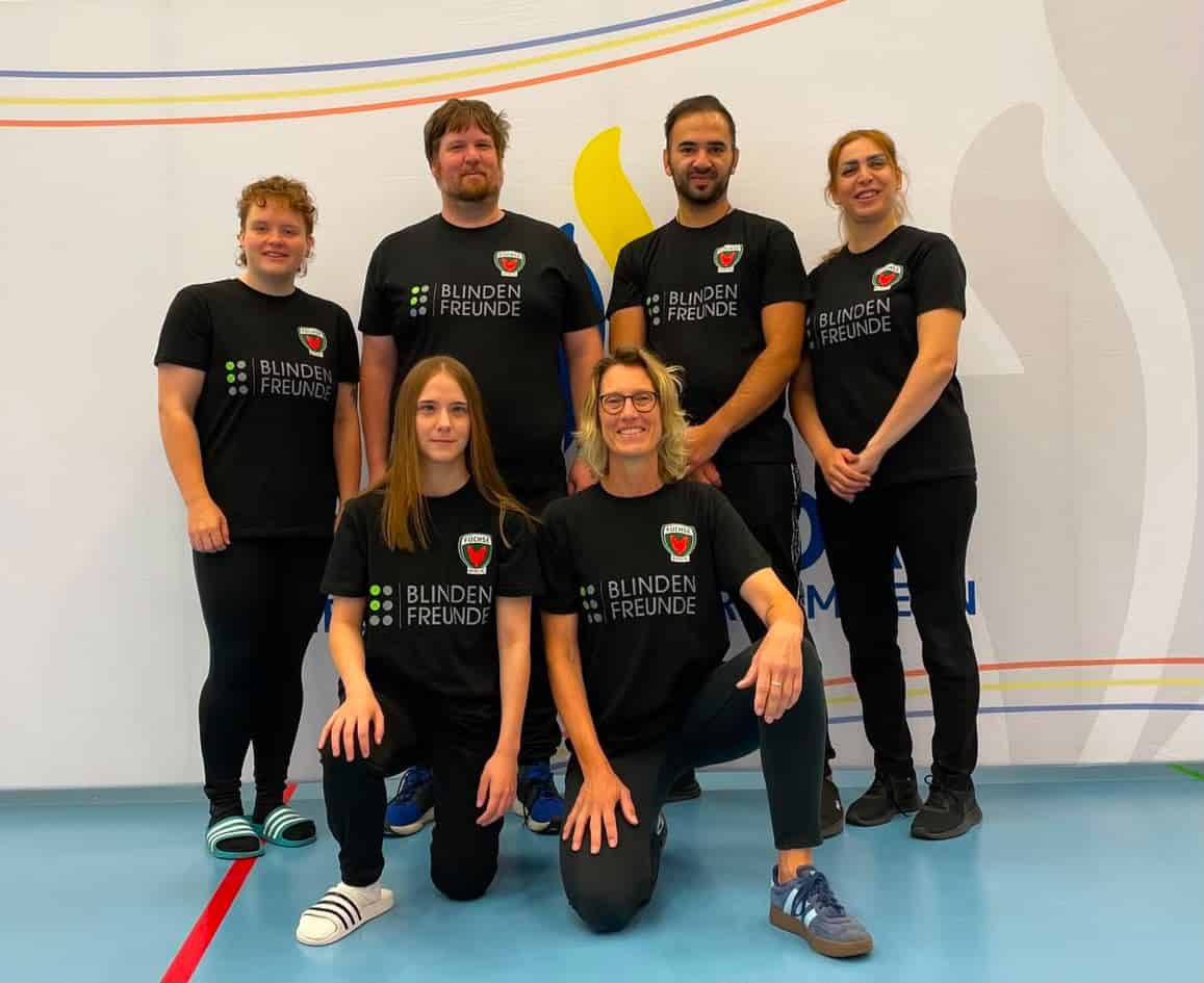 Sechs Personen in schwarzen Trikots mit dem Aufdruck Blindenfreunde stehen und knien in einer Sporthalle.