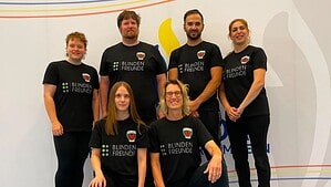 Sechs Personen in schwarzen Trikots mit dem Aufdruck Blindenfreunde stehen und knien in einer Sporthalle.