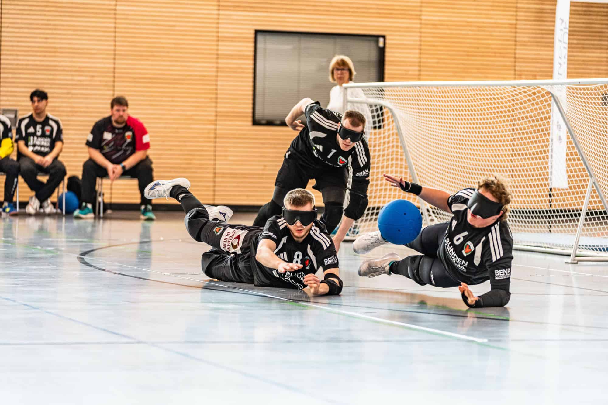 Drei Spieler eines Goalball-Teams in schwarzen Trikots mit abgedunkelten Brillen liegen und fliegen nebeneinander auf dem Hallenboden vor einem Tor, ein blauer Ball rollt auf sie zu.