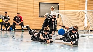 Drei Spieler eines Goalball-Teams in schwarzen Trikots mit abgedunkelten Brillen liegen und fliegen nebeneinander auf dem Hallenboden vor einem Tor, ein blauer Ball rollt auf sie zu.