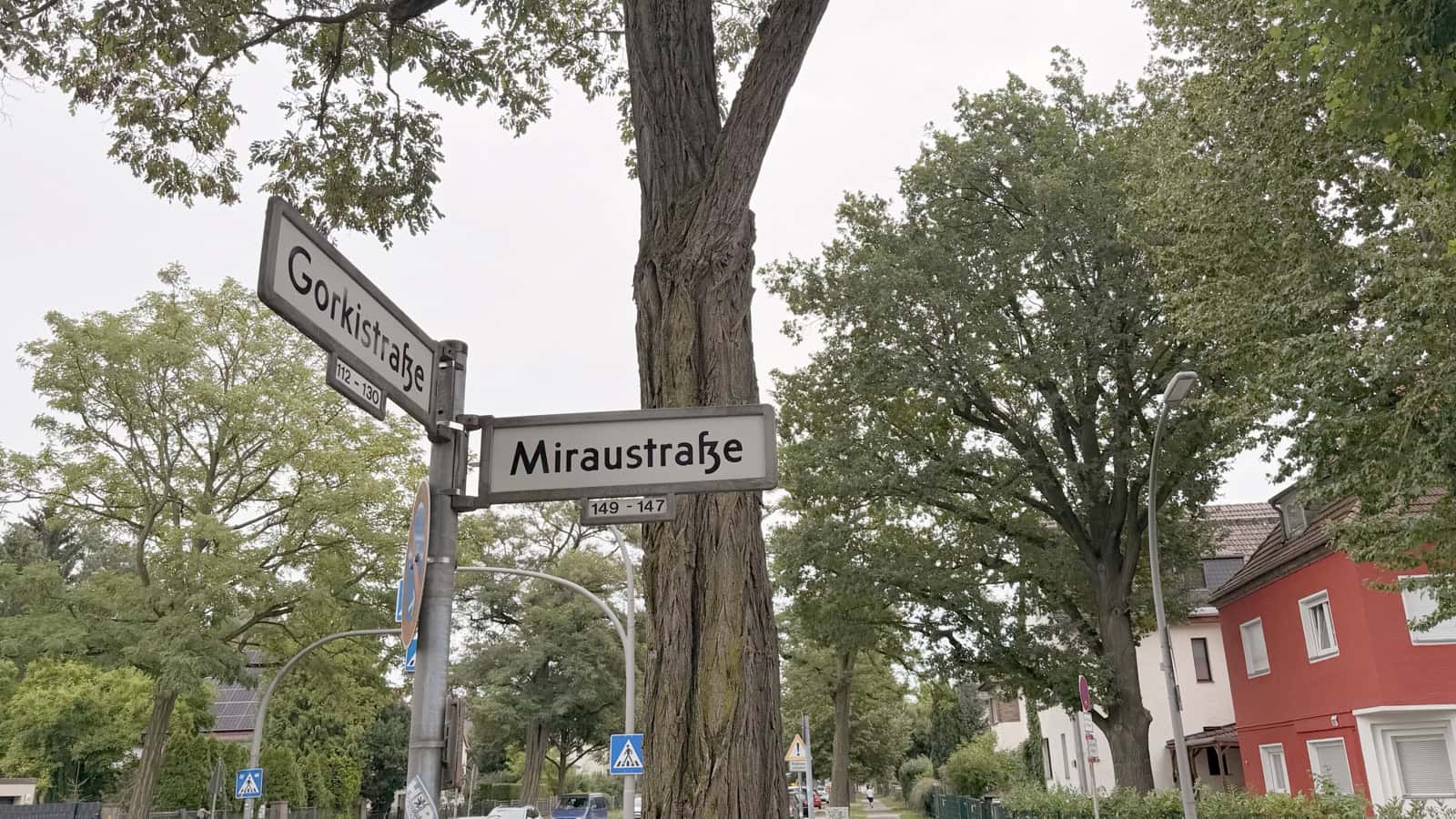 Straßenschilder mit den Namen Gorkistraße und Miraustraße an einer Kreuzung, dahinter ein alter Baum und Wohnhäuser.