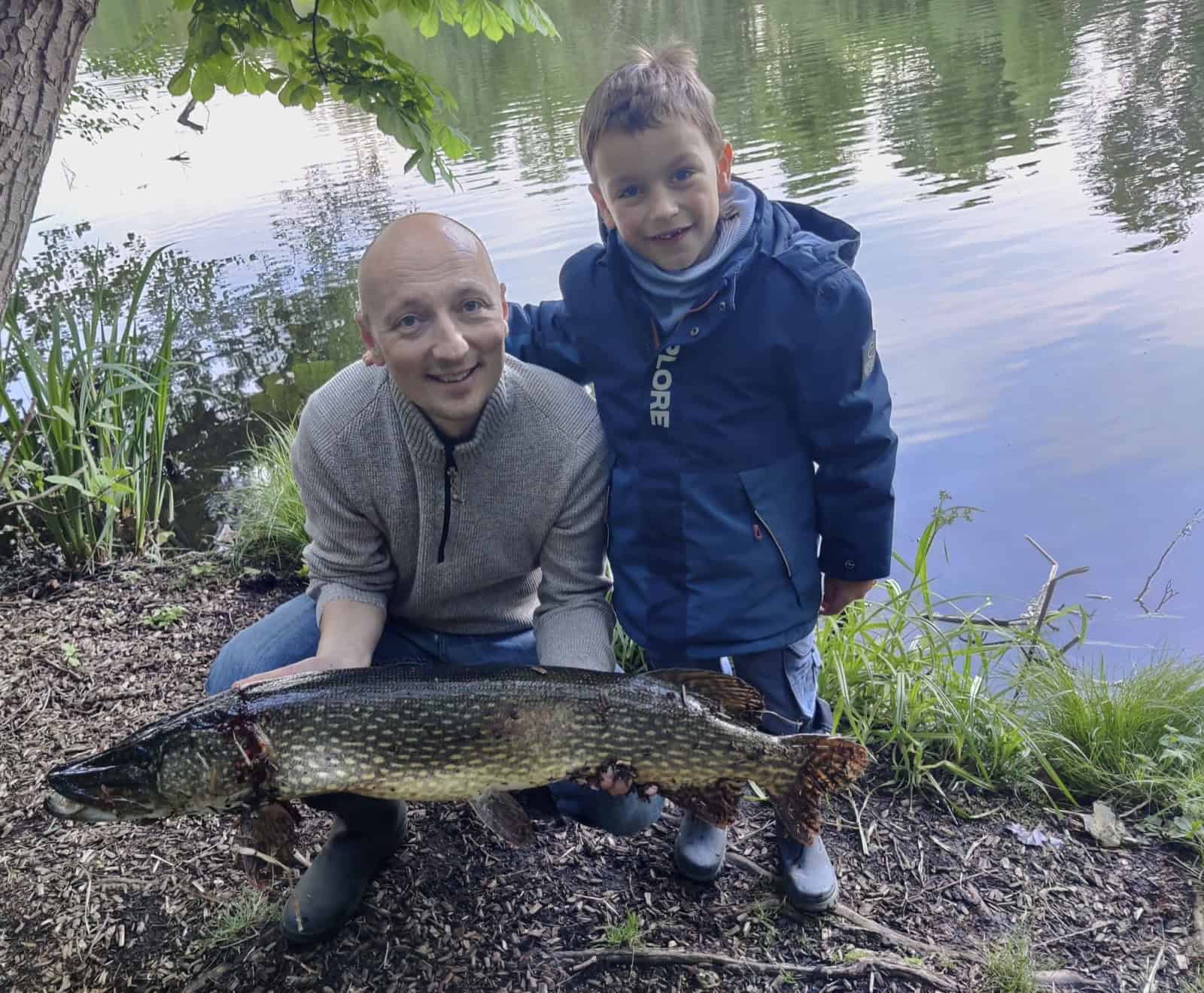 Ein Mann und ein Junge hocken am Ufer des Waldsees in Hermsdorf und halten einen großen Hecht in den Händen.