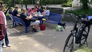 Zwei Personen im Gespräch, im Hintergrund Gäste an Tischen und auf blauen Bänken unter Bäumen