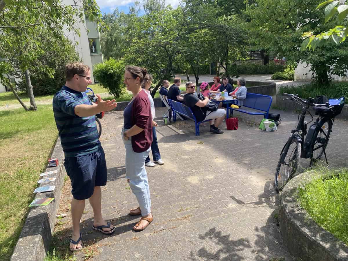 Zwei Personen im Gespräch, im Hintergrund Gäste an Tischen und auf blauen Bänken unter Bäumen