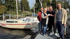 Sechs Personen stehen auf einem Bootssteg vor Segelbooten, eine Frau hält eine runde Plakette in die Kamera.