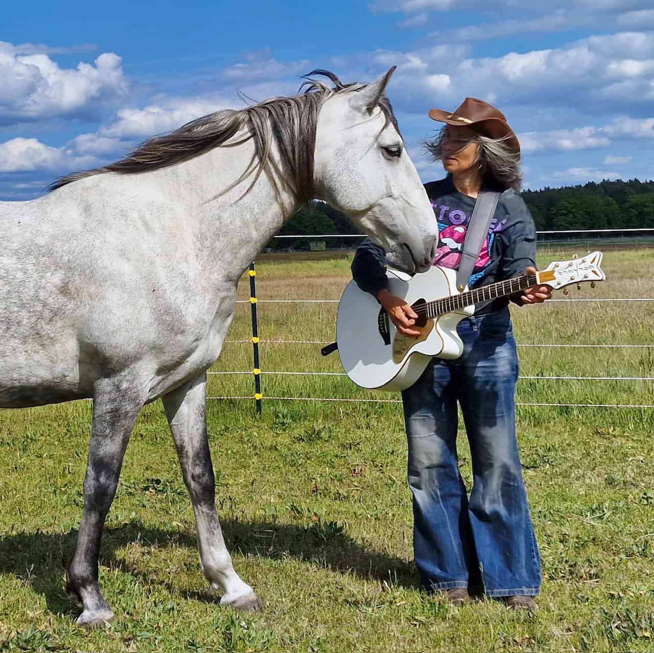 Musikerin mit Gitarre und Cowboyhut steht neben einem Schimmel auf einer Koppel