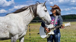 Musikerin mit Gitarre und Cowboyhut steht neben einem Schimmel auf einer Koppel