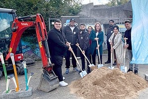 Sieben Männer und Frauen stehen mit Spaten an einem Sandhaufen neben einem kleinen Bagger, rechts eine blaue OXG-Werbefahne, im Hintergrund ein Bauzaun.