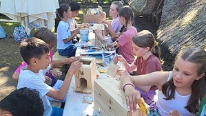 Kinder sitzen an einem langen Tisch im Freien und bauen Nistkästen aus hellem Holz, im Hintergrund ein reetgedecktes Gebäude und Bäume.