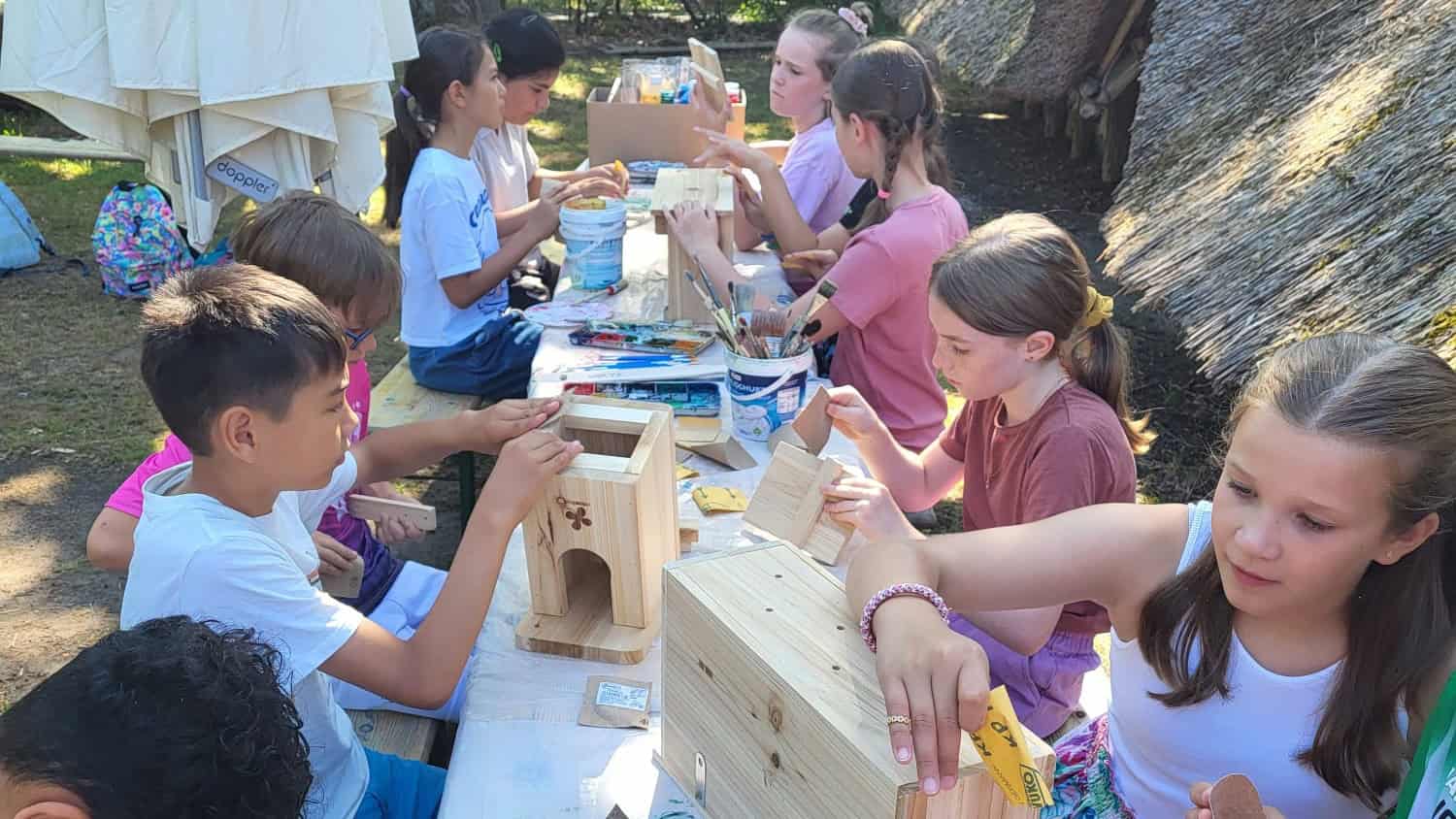 Kinder sitzen an einem langen Tisch im Freien und bauen Nistkästen aus hellem Holz, im Hintergrund ein reetgedecktes Gebäude und Bäume.