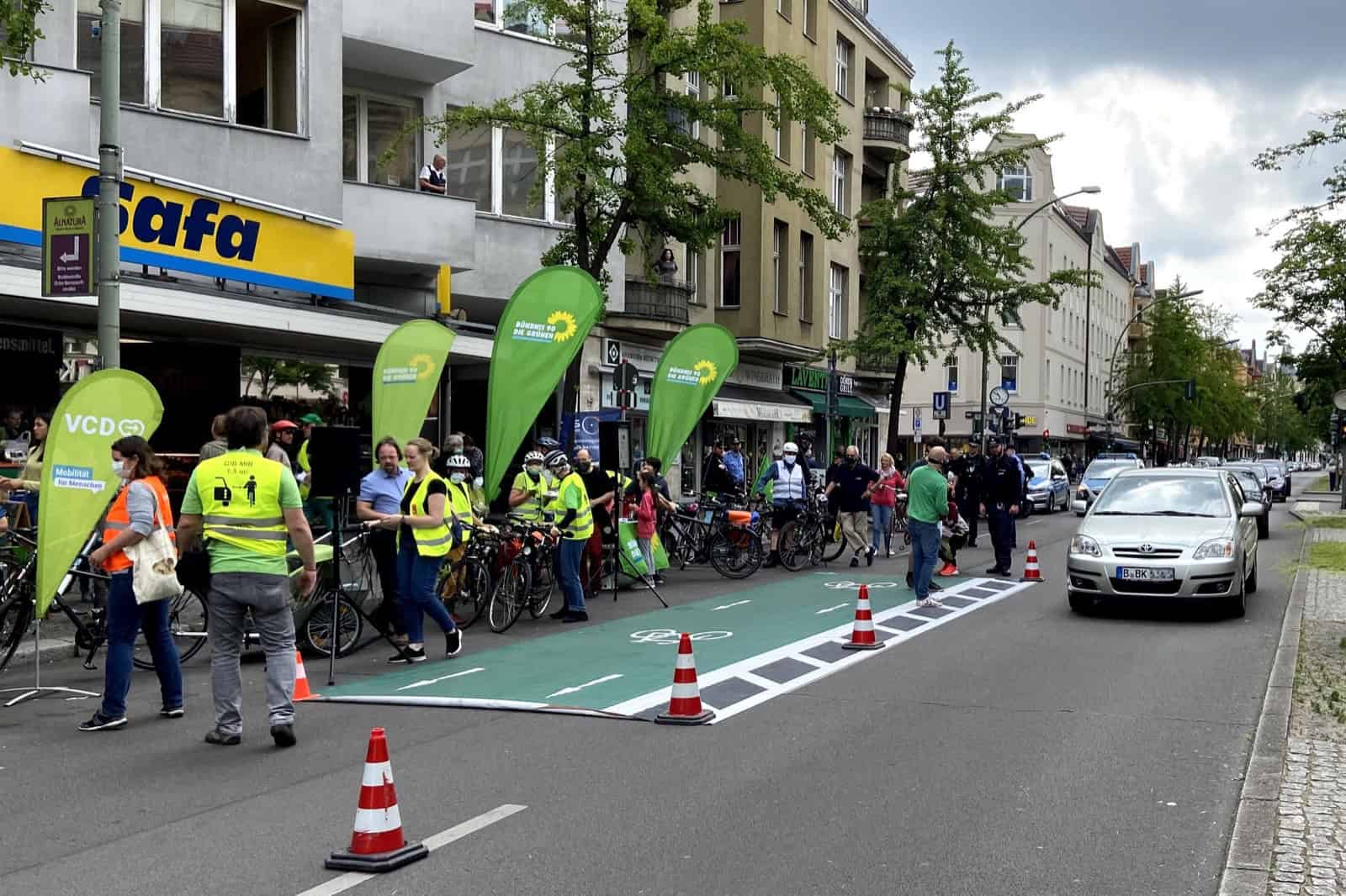 Auf der Fahrbahn der Berliner Straße liegt eine grüne Fläche mit Fahrradsymbol, abgegrenzt durch Pylonen; daneben stehen Menschen in Warnwesten mit Fahrrädern und Werbefahnen vor einer Häuserzeile mit Geschäften.