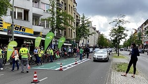 Auf der Fahrbahn der Berliner Straße liegt eine grüne Fläche mit Fahrradsymbol, abgegrenzt durch Pylonen; daneben stehen Menschen in Warnwesten mit Fahrrädern und Werbefahnen vor einer Häuserzeile mit Geschäften.